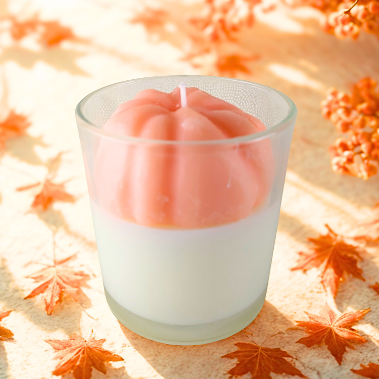 Pink pumpkin-shaped candle in a glass holder on a leaf-patterned surface